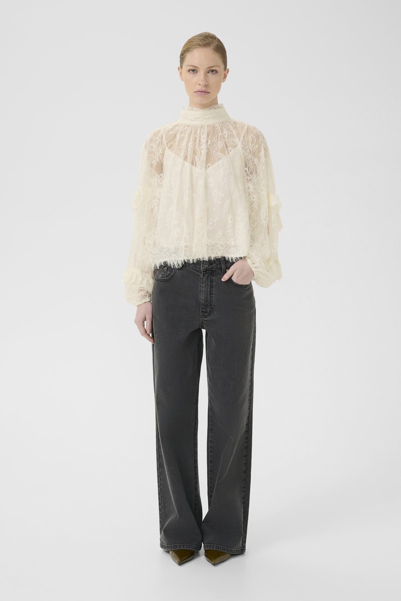 Frau trägt eine cremefarbene, transparente Spitzenbluse mit hohem Kragen und langen Ärmeln, kombiniert mit hochgeschnittenen, weit geschnittenen schwarzen Jeans und spitzen Schuhen.