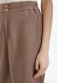 Pantalon en lin marron à coupe ample, doté d'une ceinture froncée et de deux boutons en bois pour la fermeture. Des poches latérales sont visibles.