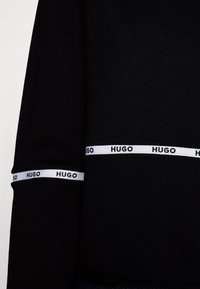 Černý rukáv a pas oděvu s bílými pruhy s opakovaným černým textovým logo "HUGO", viditelná texturovaná tkanina.