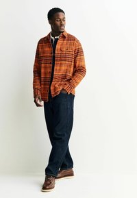 Oranjeruiten overhemd met kraag, gedragen over een donkerblauw T-shirt. Gecombineerd met donkerblauwe jeans en bruine veterschoenen.