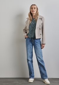 Giacca grigia chiara doppiopetto in misto lana, blusa scura a fantasia e jeans blu a zampa d'elefante, abbinati a sneakers color crema su uno sfondo semplice.