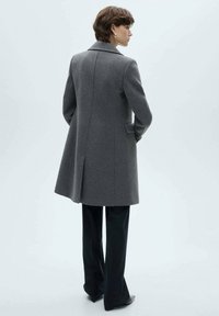 Gray wool coat with a tailored design, notched collar, and side pockets, paired with dark trousers and heels. Smooth texture and form-fitting silhouette.