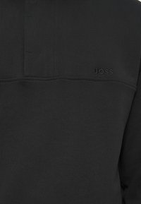 Μαύρη μπλούζα με υφή, με κολάρο που κλείνει με κουμπιά και ανάγλυφο λογότυπο "BOSS" στο αριστερό πλευρό. Λείος και απλός σχεδιασμός.