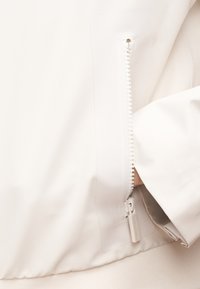 Λευκό μπουφάν με πλαϊνό φερμουάρ, διαθέτει υφασμάτινη λαβή με υφή και λείο ύφασμα. Ο σχεδιασμός έχει καθαρή, μίνιμαλ εμφάνιση.