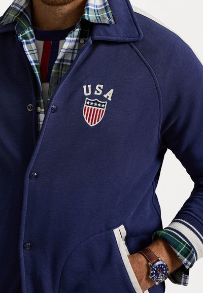 Mand iført blå jakke med "USA" skjoldemblem, lagdelt over grøn og blåternet skjorte, med et ur og hånden i lommen.