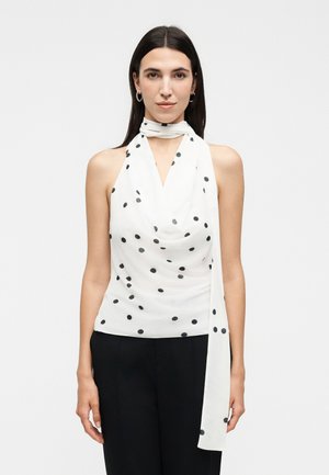 Femme aux cheveux lisses et foncés portant un haut blanc sans manches à pois avec devant drapé et une longue écharpe nouée autour du cou, pantalon noir.