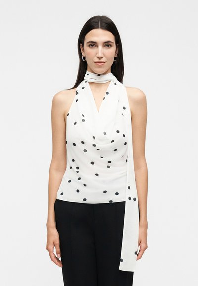 Femme aux cheveux lisses et foncés portant un haut blanc sans manches à pois avec devant drapé et une longue écharpe nouée autour du cou, pantalon noir.