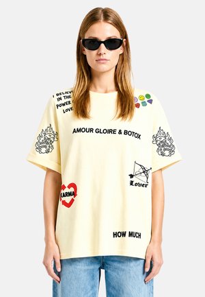 Femme portant des lunettes de soleil noires forme œil de chat, un t-shirt crème oversized avec divers textes et impressions graphiques noirs et rouges, et un jean bleu clair.