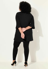 Tegola nera con maniche a svaso, abbinata a leggings neri. Modella che indossa scarpe col tacco nere, in piedi con le mani intrecciate dietro la schiena.
