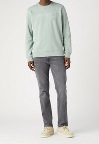 Un modello maschile indossa un maglione verde menta con la scritta "Wrangler" stampata sopra, abbinato a jeans grigi e scarpe color crema.