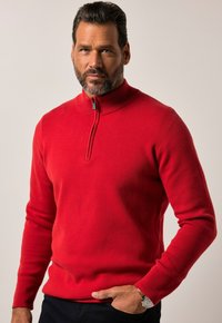 Roter gestrickter Pullover mit hohem Kragen und halbem Reißverschluss. Verfügt über lange Ärmel und eine figurbetonte Form. Glatte Textur. Über einer dunklen Hose getragen.