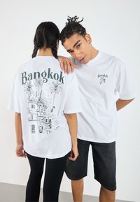 Baltos medvilnės marškinėliai su "Bangkok" tekstu priekyje ir detalia grafika gale, vaizduojančia palmės ir tuk-tuk. Įprasta fasonas.