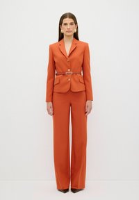 Een oranje maatpak met een fitted blazer met geknipte revers, gouden knopen en een broek met wijde pijpen. Aangevuld met een bijpassende riem.