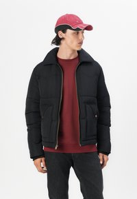 Chaqueta de plumas negra con un cuello alto, bolsillos frontales y detalles de cremallera dorada, combinada con un sudadera burdeos y una gorra roja.