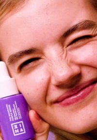 Lilla glassflaske med hvit pipette, merket "BAKUCHIOL RETINOL SERUM." Modellen holder flasken mot kinnet, smilende.