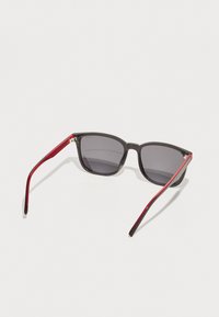 Ochelari de soare cu rame rectangulare negre și lentile gri închise. MÂinile sunt elegante, având un accent roșu lucios pe margini.