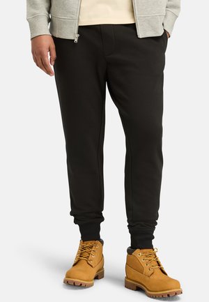 Zwarte sweatpants met geribde boorden, gecombineerd met tan werkschoenen met gele veters en gestructureerde zolen. Grijze hoodie zichtbaar.