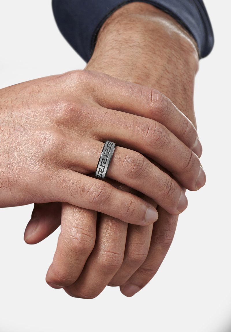 Silberner Metallring mit einem geometrischen schwarzen Muster, getragen an einer Hand mit sichtbaren Details der Hauttextur und einer weiteren Hand darunter.