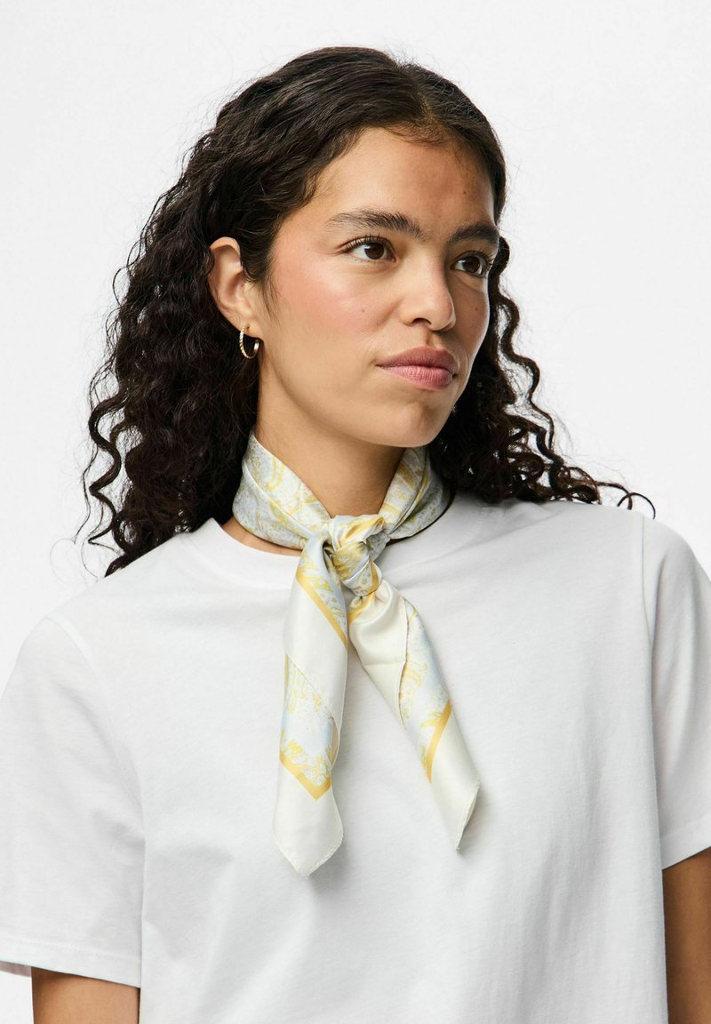 Witte zijden sjaal met een gouden en lichtblauw patroon. Om de nek geknoopt met puntige uiteinden, gecombineerd met een eenvoudige witte t-shirt.