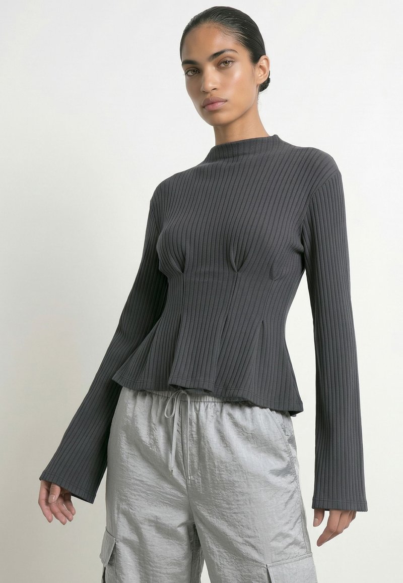 Femme aux cheveux foncés tirés en arrière, portant un haut côtelé ajusté à manches longues gris foncé et un pantalon gris clair à cordon avec poches latérales.