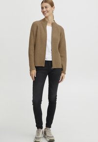 Brown ribbed knit cardigan with a stand collar and open front, worn over a white shirt, paired with black skinny pants and light-colored sneakers.