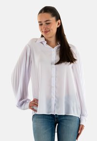 Camicia bianca con tessuto liscio e setoso, caratterizzata da chiusura frontale con bottoni, collo classico e maniche a sbuffo plissettate. Abbinata a denim.