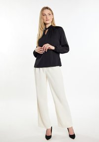 Schwarze Bluse mit langen Ärmeln und Ausschnit in Form eines Schlüssellochs, kombiniert mit weißen, weit geschnittenen Hosen und schwarzen High Heels. Glatte Stoffoberfläche.