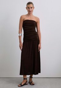 Vestido marrón sin mangas con textura arrugada, silueta fluida y falda larga. Combinado con sandalias negras con tachuelas y un brazalete plateado.