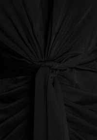 Tissu noir soigneusement rassemblé et noué en un nœud, créant des plis et un drapé texturé autour du nœud central.