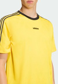 Žlté tričko Adidas s čiernym logom, okrúhlym výstrihom s čiernym lemom a dvojitými čiernymi pruhmi na ramenách; hladká textúra.