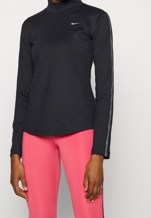 Personne portant un haut Nike noir à manches longues avec un col haut et des leggings roses, debout devant un fond clair uni.