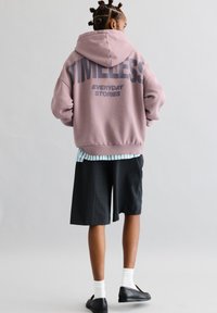 Preveliki ružičasti hoodie s grafikom "VJEČNE SVAKODNEVNE PRIČE" u tamno sivoj, uparen s crnim kratkim hlačama i bijelim čarapama. Glatka tekstura.