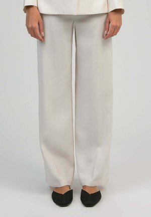 Femme portant un pantalon beige à jambes larges et des chaussures plates noires à bout pointu, debout devant un fond uni gris clair.