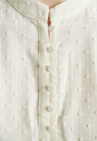 Gros plan d'un chemisier blanc texturé avec de petits motifs en losange et des boutons recouverts de tissu à l'avant.