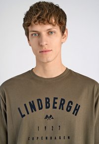 Camiseta de manga larga verde oliva hecha de algodón suave. Presenta "LINDBERGH 1927 COPENHAGUE" impreso en texto negrita azul marino en el pecho.
