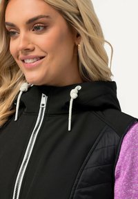 Gilet nero con chiusura a zip, caratterizzato da un cappuccio e corde bianche. I lati presentano una texture trapuntata, abbinata a un materiale viola.