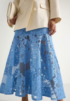 Person iført en cremefarvet blazer og holder en cremefarvet clutch, med et knælængde blåt blomstret blonder nederdel mod en ensfarvet baggrund.