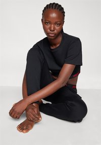 Ensemble t-shirt et pantalon en coton noir, avec un design court. Le t-shirt a un col rond ; le pantalon présente un logo rouge "HUGO" sur la ceinture.