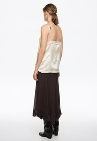 Kobieta z niskim kucykiem, ubrana w kremową satynową koszulkę na ramiączkach, ciemnobrązową spódnicę z koronkowym dołem i czarne kozaki za kolano, zwrócona tyłem.