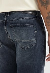Donkerblauwe denim jeans met een rechte snit, twee achterzakken en een leren label met "PURE PATH" op de tailleband.