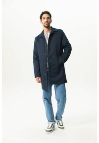 Marineblauer, mittellanger Mantel aus strapazierfähigem Material mit geradem Schnitt, vorderen Knöpfen und seitlichen Taschen, getragen über einem gestreiften Hemd und Jeans.