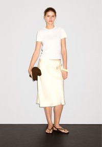 Bílý přiléhavý tričko, saténová krémová midi sukně s elastickým pasem, hnědá texturovaná kabelka a hnědé sandály na pásky na tmavé podlaze.