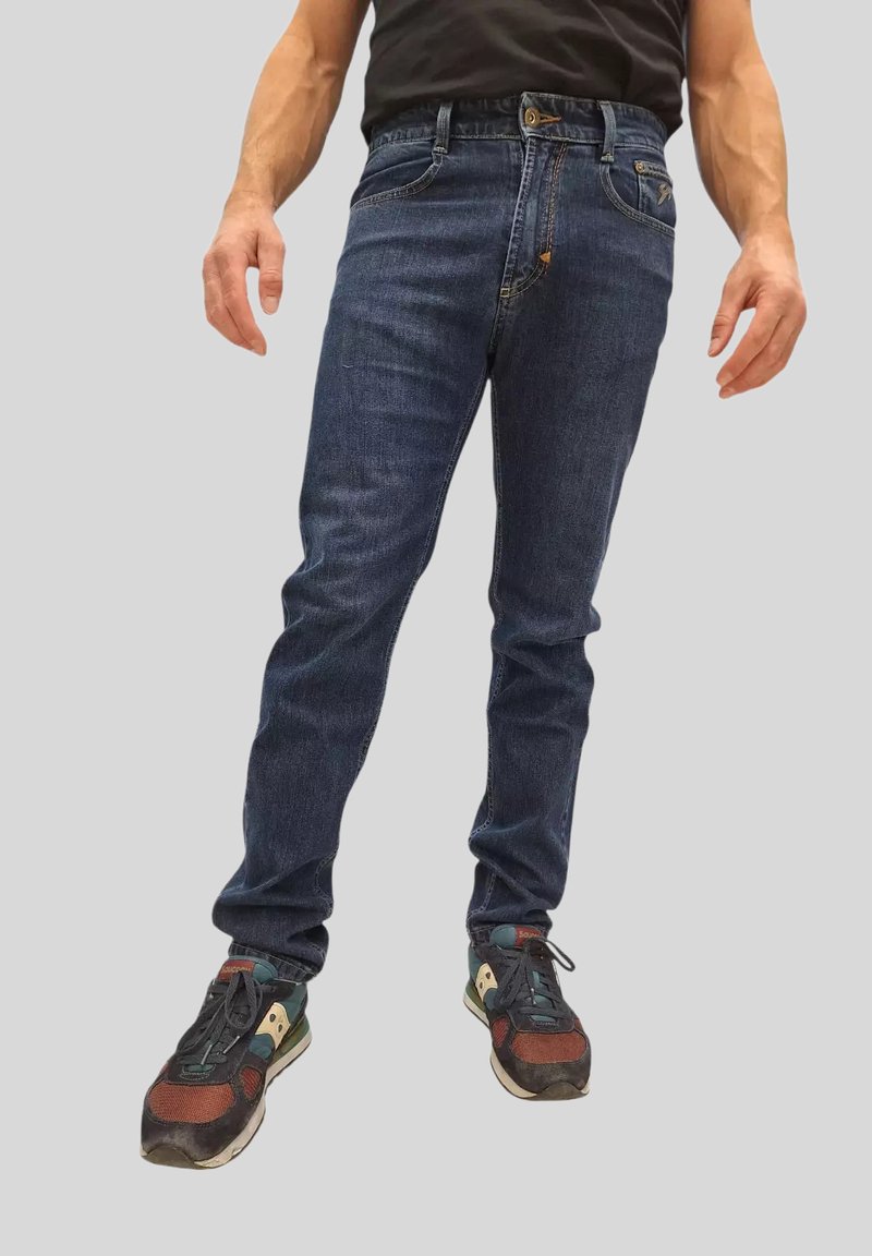 Jeans in denim blu scuro con vestibilità slim, design a cinque tasche e dettagli di cucitura a contrasto, indossati con scarpe da ginnastica multicolori.