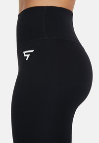 Čierne legíny s vysokým pásom vyrobené z elastického materiálu, s bezšvovým dizajnom a malým bielym logom na stehne.