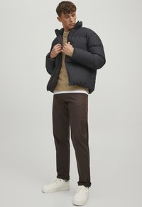 Schwarze Daunenjacke mit hohem Kragen, leichter Polsterung und Reißverschluss, getragen über einem beigen Strickpullover und braunen Hosen; weiße Turnschuhe.