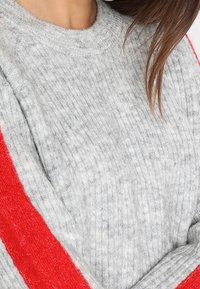 Pullover grigio a maglia con texture a costine, scollatura tonda e una vistosa striscia rossa che corre diagonalmente lungo la manica.
