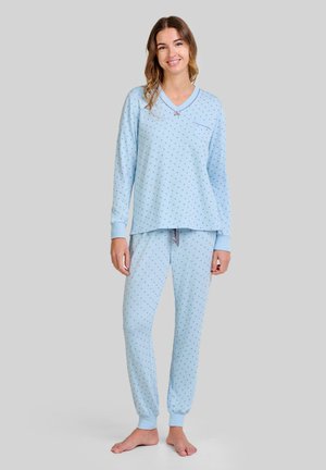Mujer de pie descalza, vestida con un conjunto de pijama de manga larga azul claro con pequeños lunares oscuros, frente a un fondo gris liso.