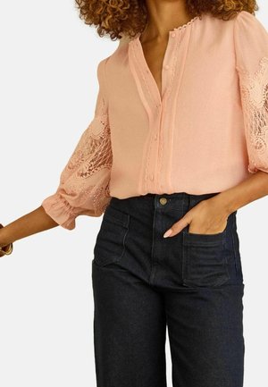 Frau trägt eine hellrosa Bluse mit Spitzen-Ballonärmeln und hoch taillierte dunkle Jeans mit vorderen aufgesetzten Taschen, Hand in der Tasche.