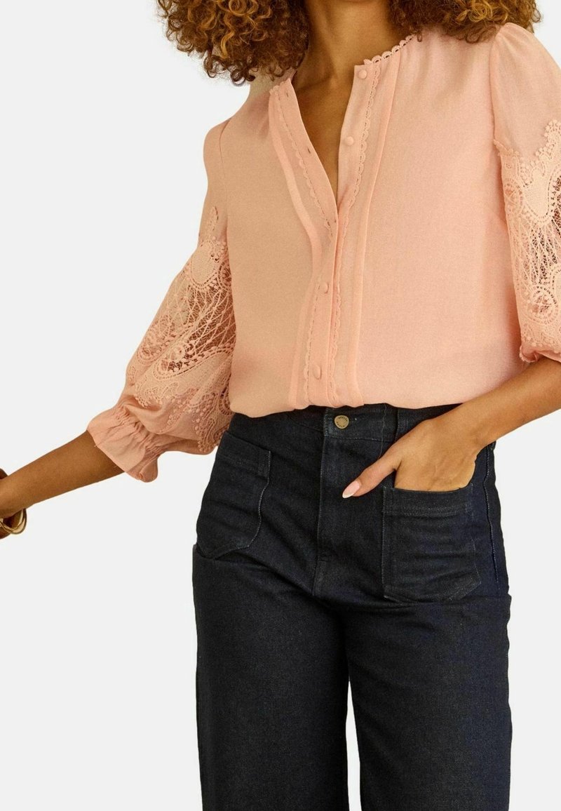 Femme portant un chemisier rose clair avec manches ballon en dentelle et un jean foncé taille haute avec des poches plaquées à l’avant, main dans la poche.