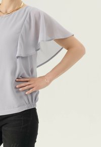Donna che indossa una blusa lilla chiaro con maniche a pipistrello, pantaloni neri, un bracciale dorato e collane dorate a strati, mentre posa con una mano sulla vita.